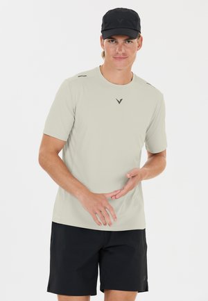 Young man wearing a light beige short-sleeve shirt, black shorts, and a black cap, standing against a white background.