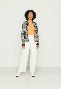 Casaco bombardeiro preto com estampa de folha de palmeira branca, top laranja com abertura em forma de olho, jeans brancos de cintura alta e ténis brancos. Óculos de sol redondos.