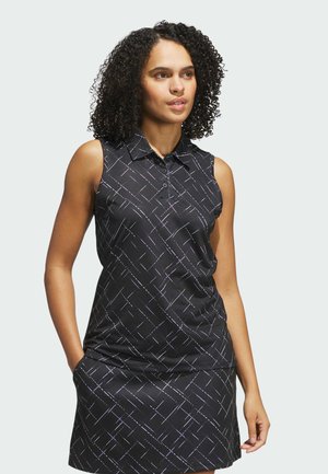 Kvinde med krøllet hår iført en ærmeløs sort kjole med krave og hvidt geometrisk linjemønster, stående med hænderne i lommerne og kiggede mod højre.