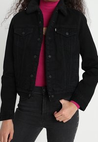 Czarna dżinsowa kurtka z kołnierzem podszytym polarem, zapinana na guziki, z kieszeniami na piersiach. Nosi ją na różowej, prążkowanej bluzce i zestawia z ciemnymi dżinsami.