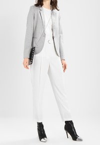 Šedý blazer přes bílý tričko, v kombinaci s bílými pruhovanými kalhotami a černými kotníkovými botami s lesklými stříbrnými akcenty. Kabelka s řetízkovým držadlem.