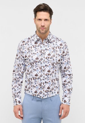 Mand iført hvid skjorte med blomstertryk og lyseblå bukser, stående mod en ensfarvet baggrund.