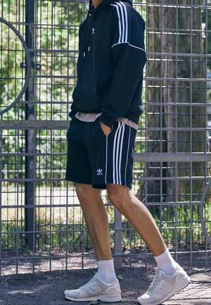 Osoba ubrana w czarną bluzę z kapturem Adidas, pasujące szorty z białymi paskami, białe trampki, stojąca obok metalowego ogrodzenia na zewnątrz.