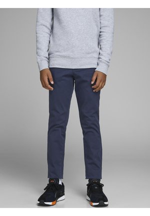 Persona con sudadera gris, pantalones azul marino y zapatillas negras con suelas blancas y detalles naranjas, de pie contra un fondo gris liso.
