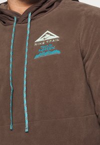Felpa con cappuccio marrone in pile con lacci teal e grafiche ricamate, tra cui "Nike Trail" e "Tour du Mont Blanc." Texture morbida.