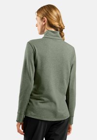 Frau mit rötlichem Haar in einem Zopf, die einen langärmligen, olivgrünen Rollkragenpullover und schwarze Hosen trägt, mit dem Rücken zu einer weißen Hintergrund.