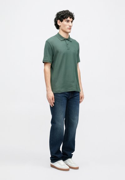 Polo vert en coton, avec un col et des manches courtes, associé à un jean bleu foncé et des baskets blanches.