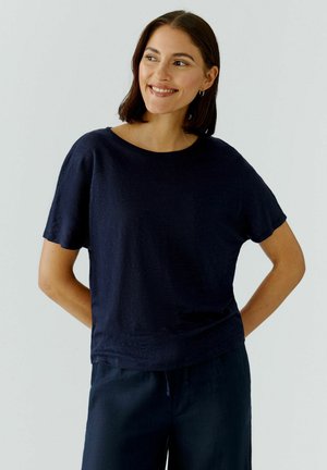 Femme aux cheveux mi-longs foncés, vêtue d'un haut bleu marine à manches courtes et d'un pantalon assorti, souriante et regardant vers la droite.