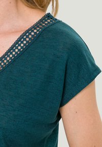 Femme portant un haut à manches courtes teal avec une bordure en treillis décoratif le long du décolleté en V, visible de l'épaule au haut de la poitrine.