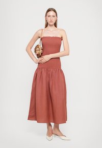 Strapless, rust-colored dress with smocked bodice and a flowing, gathered skirt. Paired with a woven circular handbag and white pointed shoes.
