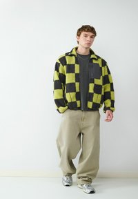 Geruite jacket in zwart en neon groen, van fleecemateriaal met een ritszak. Gecombineerd met lichtgekleurde baggy broek en zilveren sneakers.