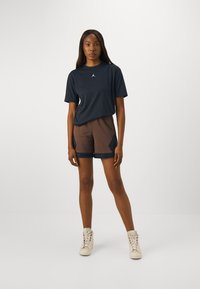 T-shirt marine à manches courtes avec logo blanc ; short marron avec accents noirs et motif en losanges ; associé à des baskets montantes beiges.