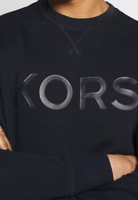 Persona che indossa una felpa nera con la scritta "KORS" in rilievo in grandi lettere sul petto, con le braccia incrociate in vita.