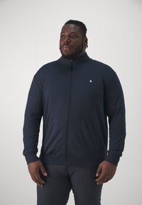 Mornarska zip-up dukserica izrađena od mekanog materijala, s visokim ovratnikom, dva prednja džepa i malim logotipom na prsima. Glatka tekstura.