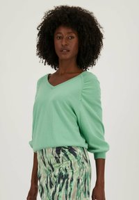 Licht mintgroen sweater met een V-hals en gerimpelde details op de schouders, gecombineerd met een rok met een kleurrijk patroon in groene en zwarte tinten.