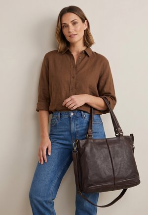 Kobieta ubrana w brązową koszulę na guziki i niebieskie dżinsy trzyma dużą, ciemnobrązową skórzaną torbę typu tote za uchwyty.