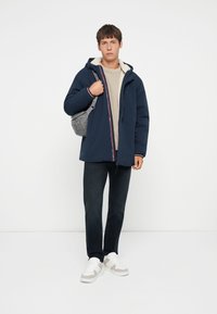 Giacca a vento blu navy con fodera interna color crema, indossata sopra un maglione beige, abbinata a jeans scuri e sneaker bianche.