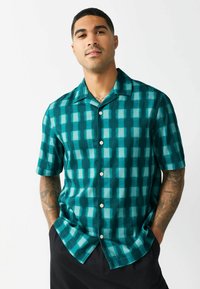 Cămașă cu mânecă scurtă, de culoare verde, cu motive în carouri, cu închidere cu nasturi și guler. Modelul prezintă nuanțe variate de teal și albastru deschis.