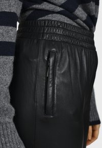 Pantaloni in pelle nera con vita elasticizzata, dotati di una tasca con zip laterale. Il materiale ha una texture liscia e un design elegante.