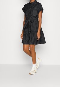 Dark denim shirt dress with short sleeves, two chest pockets, a tied waist, and a tiered skirt. Paired with white and tan sneakers.