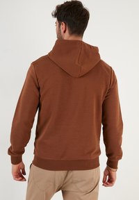Sudadera marrón con un diseño clásico, cuenta con un bolsillo tipo canguro y puños acanalados, fabricada en suave tejido de algodón con un acabado texturizado.