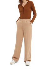 Kvinde iført en brun langærmet henley-skjorte, beige vide bukser, hvide sneakers og et armbånd, stående med hænderne i lommerne.