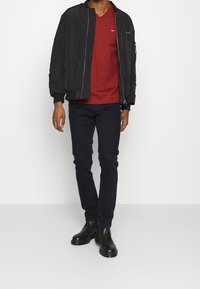 Herroutfit med en svart bomberjacka, röd v-ringad t-shirt, mörka jeans och svarta ankelboots; slät tyg och åtsittande design.