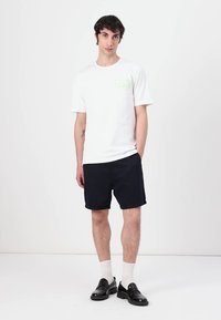 Hvit bomull t-skjorte med en grønn logo på brystet, kombinert med marineblå shorts. Modell iført svarte sko og hvite sokker, stående mot en ensfarget bakgrunn.