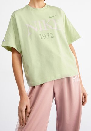 Osoba ubrana w jasnografitowy T-shirt Nike z napisem „1972” oraz różowe spodnie Nike Air z białym logo na lewej nogawce.