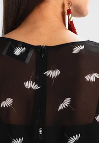 Blusa negra con un tejido transparente y patrones florales blancos. Incluye un cierre en la parte trasera y pendientes con borlas visibles en el modelo.