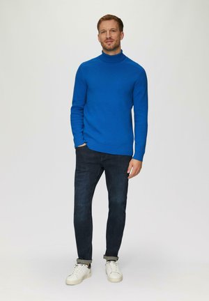 Blauwe coltrui in een gebreide stof, gecombineerd met donkerblauwe jeans en witte sneakers. De trui heeft geribbelde manchetten en een boord.
