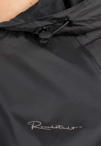 Giacca nera impermeabile con fodera in rete, cappuccio regolabile e chiusura con passante; presenta un logo ricamato bianco sul davanti.