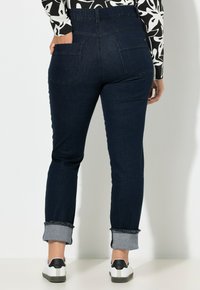 Jeans de mezclilla azul oscuro con corte ajustado, con puños enrollados, dos bolsillos traseros y una cinturilla de altura media. Combinados con zapatillas blancas.