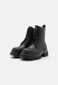 Botas negras hasta el tobillo con suelas gruesas, de cuero sintético suave, puntas redondas y diseño de cordones. Presenta una lengüeta y una marca en el lateral.