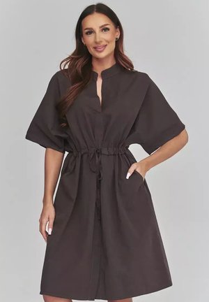 Kobieta z długimi brązowymi włosami w czarnej sukience do kolan z szerokimi rękawami i ściągaczem w talii, stojąca z jedną ręką w kieszeni.