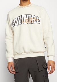 Cream-colored sweatshirt with a ribbed collar, featuring bold "COUTURE" text in contrasting colors and a relaxed fit. Smooth texture, long sleeves.