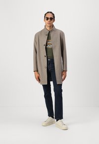 Μπεζ παλτό από μάλλινο υλικό με όρθιο γιακά και κλείσιμο με κουμπιά, φορεμένο πάνω από ένα πράσινο μπλουζάκι και σκούρα τζιν, συνδυασμένο με κρέμ πάνινα παπούτσια.