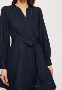 Abito blu navy con bottoni, maniche lunghe, cintura in vita e dettagli a taglio lungo le cuciture e l'orlo, caratterizzato da una vestibilità comoda e una texture morbida.