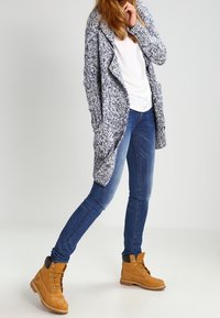 Stickad kofta i marinblått och vitt, tillsammans med en vit skjorta och blå jeans. Bruna snörskor med en stabil sula och guldfärgade öglor.