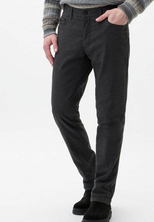 Mężczyzna w ciemnoszarych, dopasowanych spodniach, szarym swetrze w paski i czarnych butach, z jedną ręką w kieszeni, stojący na tle białego tła.