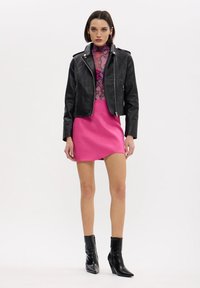 Donna che indossa una giacca di pelle nera, un top con motivo in pelle di serpente rosa, una gonna rosa brillante e stivali neri alla caviglia, in piedi contro uno sfondo bianco semplice.