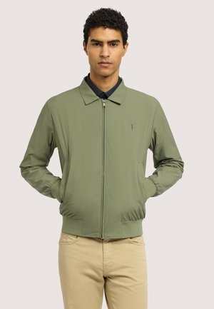 Mand iført olivengrøn jakke med lynlås og krave samt lyse beige bukser, stående med hænderne i lommerne mod en ensfarvet baggrund.