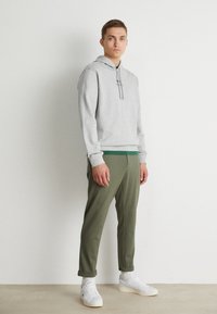 Giovane uomo in piedi all'interno che indossa un hoodie grigio chiaro, pantaloni verde oliva, calzini bianchi e sneakers bianche davanti a un muro bianco semplice.