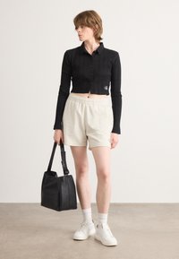 Svart cropped langermet skjorte med vertikale riller og knapplukking, sammen med lysebeige shorts med elastisk liv og en svart totebag.