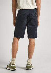 Hombre vestido con pantalones cortos negros hasta la rodilla, una camiseta de rayas beige, calcetines beige y zapatillas verdes oliva, de pie contra un fondo liso.