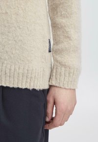 Suéter de punto beige hecho de una tela suave y texturada. Cuenta con puños y dobladillo de canalé. Etiqueta visible en el lateral. Pantalones oscuros mostrados parcialmente.
