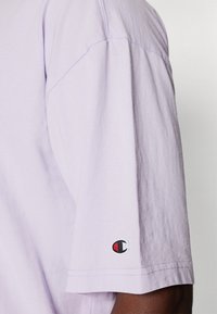 Lys lilla kortærmet t-shirt lavet af bomuld. Har en syet søm og et lille broderet logo i rød, sort og hvid påærmet.