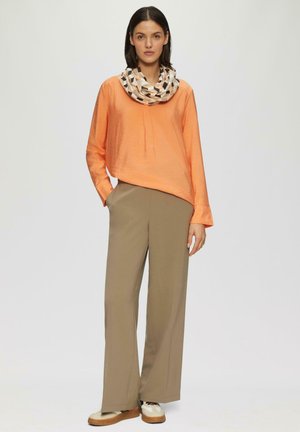 Femme debout portant un haut à manches longues orange, un pantalon large beige, une écharpe à motifs et des chaussures compensées beiges sur un fond uni.