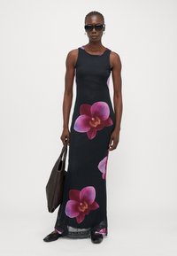 Robe maxi en tissu noir avec de grandes impressions d'orchidées roses. Modèle sans manches avec une silhouette ajustée. Le mannequin porte un sac tote marron.