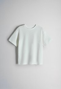 Camiseta blanca lisa de manga corta colgada contra una pared gris clara, con cuello redondo y corte holgado.